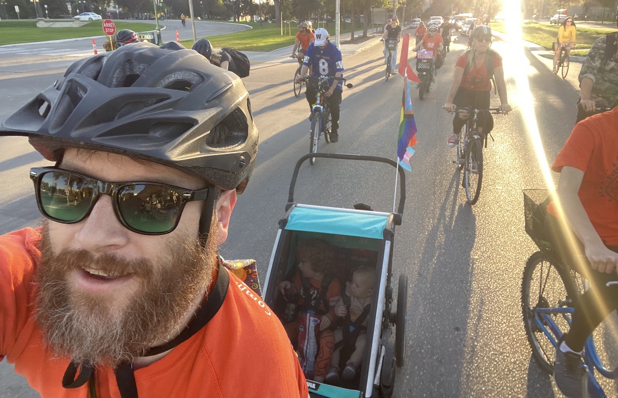 Andrew Kohan, a white man with a beard and wearing an orange shirt and a bike helmet, rides in a group bike ride with a trailer with two children and pride flags