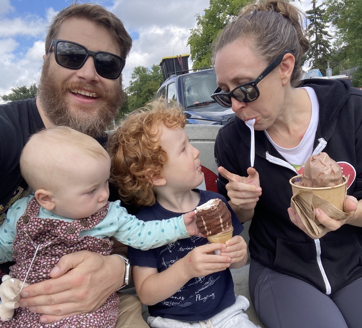 andrew Kohan, a white man with a beard, holding a baby who is grabbing the ice cream in the cone help by another child, sitting next to a woman eating ice cream with a spoon